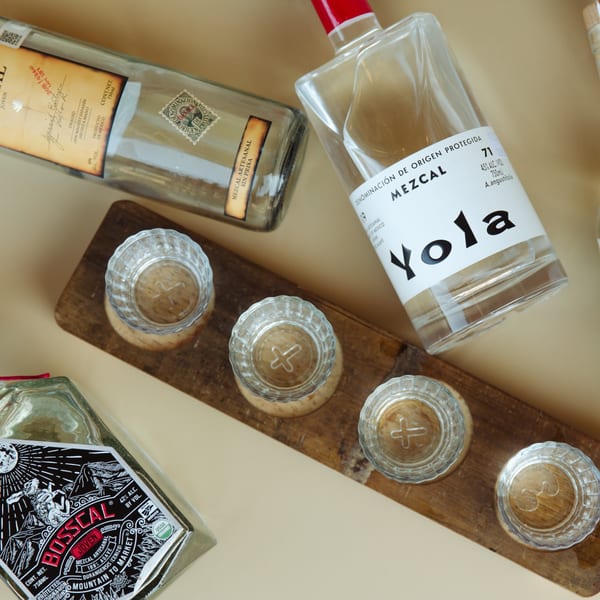 Three bottles of mezcal and four empty shot glasses on a wooden tray.