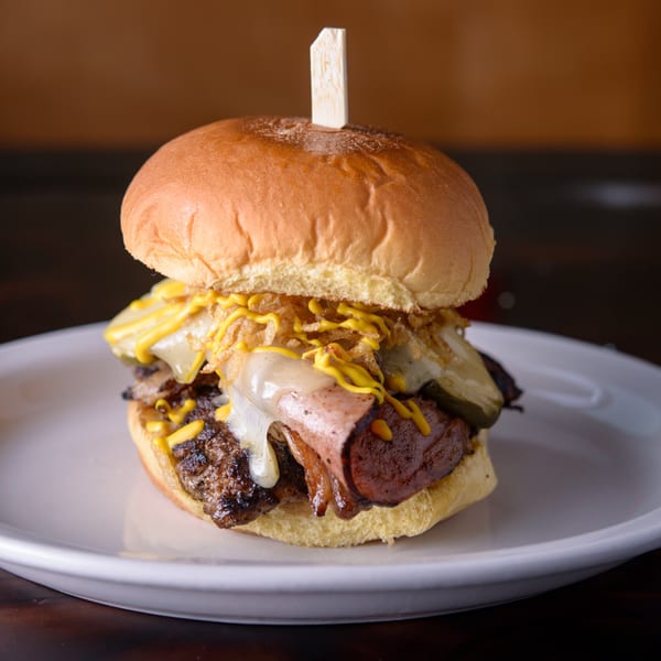 Cuban Burger with melted cheese, crispy onions, mustard, and a wooden skewer on a white plate.