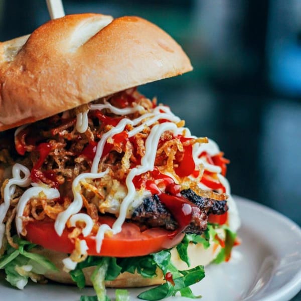 A Cuban Burger with a toasted bun, crispy onions, lettuce, tomato, and sauces on a white plate.