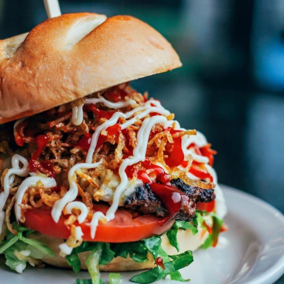 A Cuban Burger with a toasted bun, crispy onions, lettuce, tomato, and sauces on a white plate.