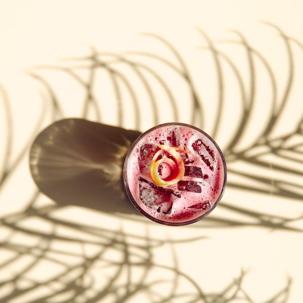 Overhead view of a vibrant red Cuban cocktail with ice and a citrus peel, casting palm leaf shadows.
