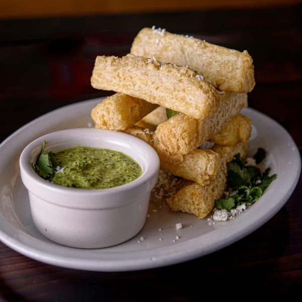 A plate of golden-brown Cuban yuca fries, sprinkled with cheese, next to a white bowl of green cilantro sauce.