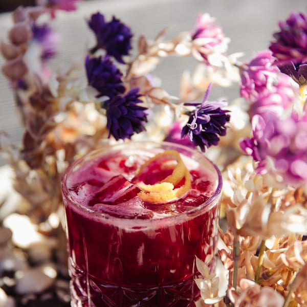 A vibrant red Cuban cocktail with ice and an orange peel garnish, surrounded by purple and beige flowers.