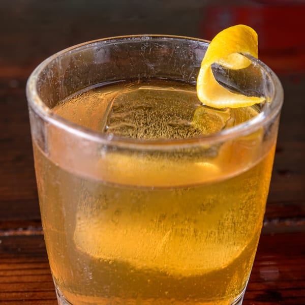 Close-up of a golden cocktail with ice and a lemon peel garnish on a dark wooden surface.